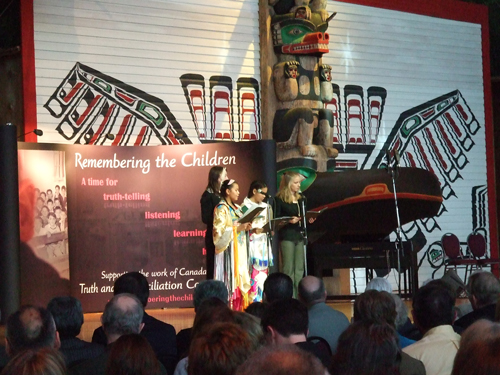 Groupe de quatre personnes, incluant des enfants, dont deux en tenues traditionnelles, debout sur une scène devant un mât totémique et un panneau indiquant Remembering the Children (« se souvenir des enfants »).