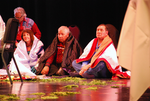Trois personnes autochtones sont assises sur une scène, enveloppées dans des couvertures, avec des branches de cèdre devant elles.