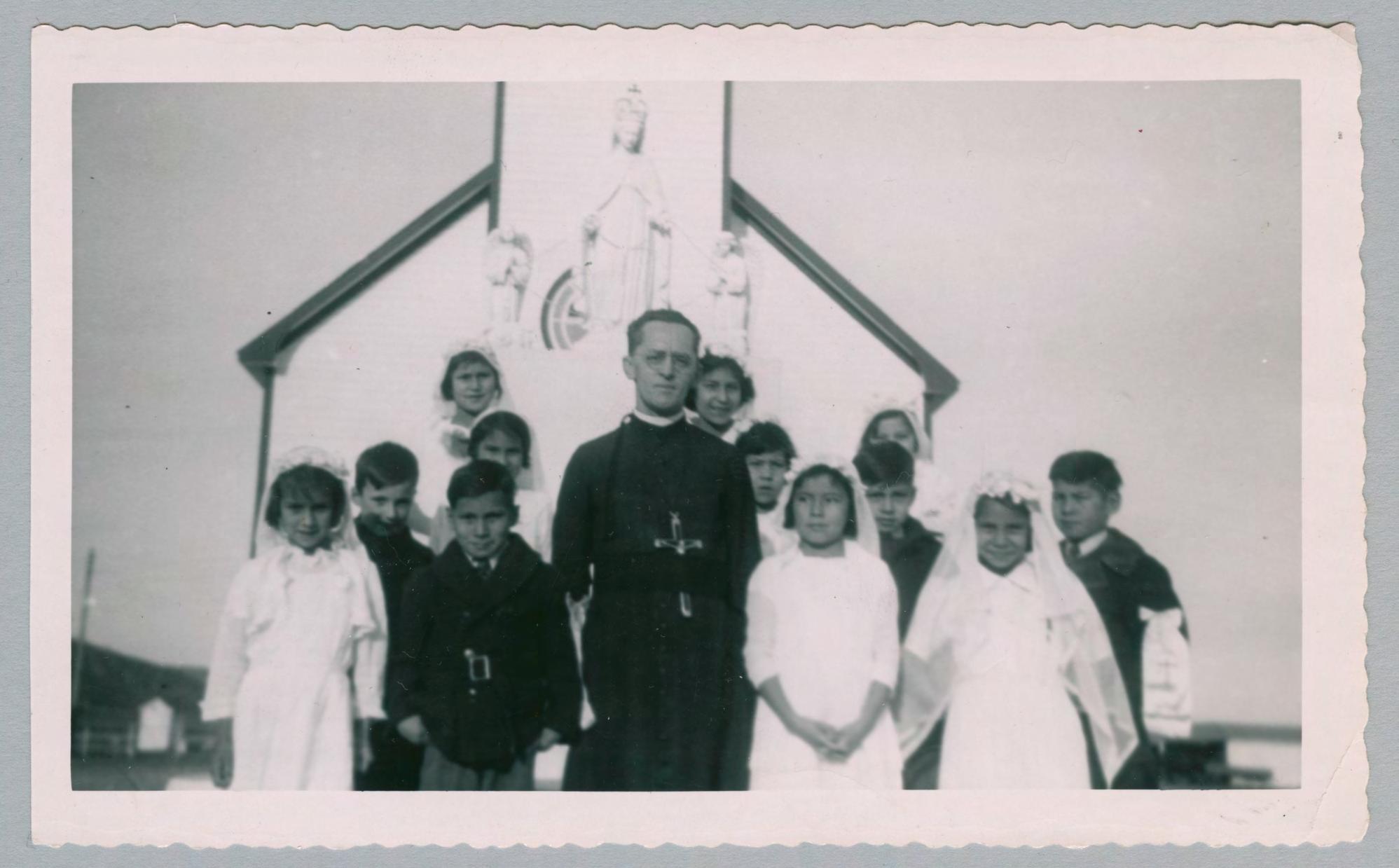 Père Carriere stands in front of a church surrounded by children. The boys are in black suits and the girls are in white dresses and veils.