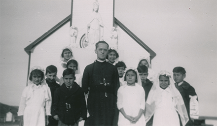 Père Carriere stands in front of a church surrounded by children. The boys are in black suits and the girls are in white dresses and veils.