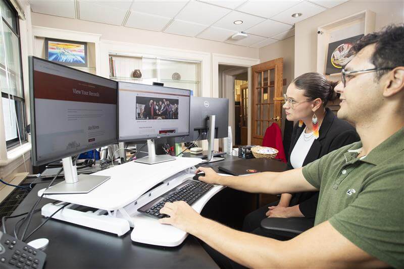 Two people sit at a desk, looking at a computer.