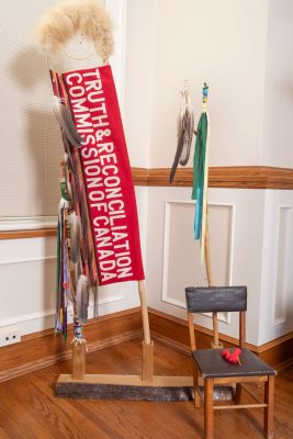 A ceremonial Walking Stick with beaded university crests, symbolizing reconciliation commitments, alongside two Eagle Staffs: one commissioned for the Truth and Reconciliation Commission of Canada and one gifted by Reconciliation Walkers, representing healing and awareness.