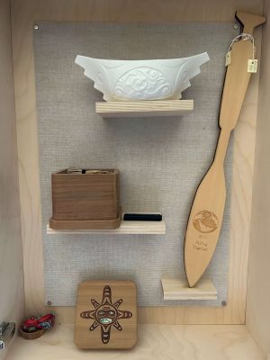 A collection of three artifacts including 'Spirit Canoe III,' a porcelain canoe by Terry Jackson; a reconciliation paddle from the Pulling Together Canoe Society; a wooden box with braided sweetgrass and a USB  and a mini bentwood box by Shane Jackson featuring a sun mask, which once held documents for missing children research.