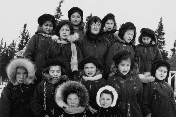 Black and white photo of Indigenous children and an adult woman in winter clothing, standing in a snowy landscape.