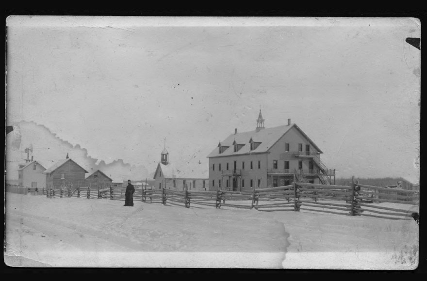 Winter scene of a 19th-century settlement: a large two-story building, church steeple, split-rail fence, and a solitary figure in the snow.